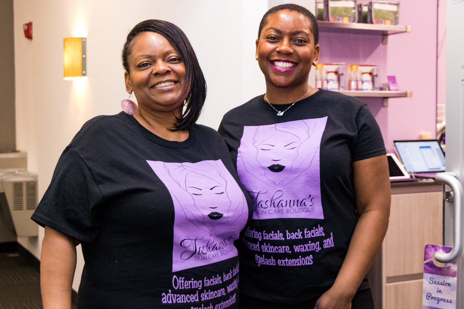 Two women wearing black t-shirts with a purple logo and text in a salon setting.