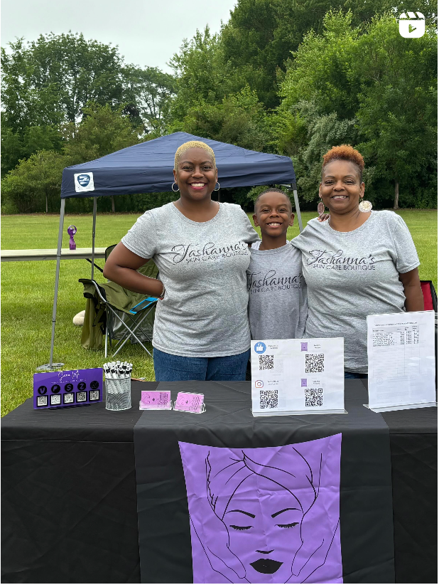 Three people standing behind a table with products and promotional materials outdoors.